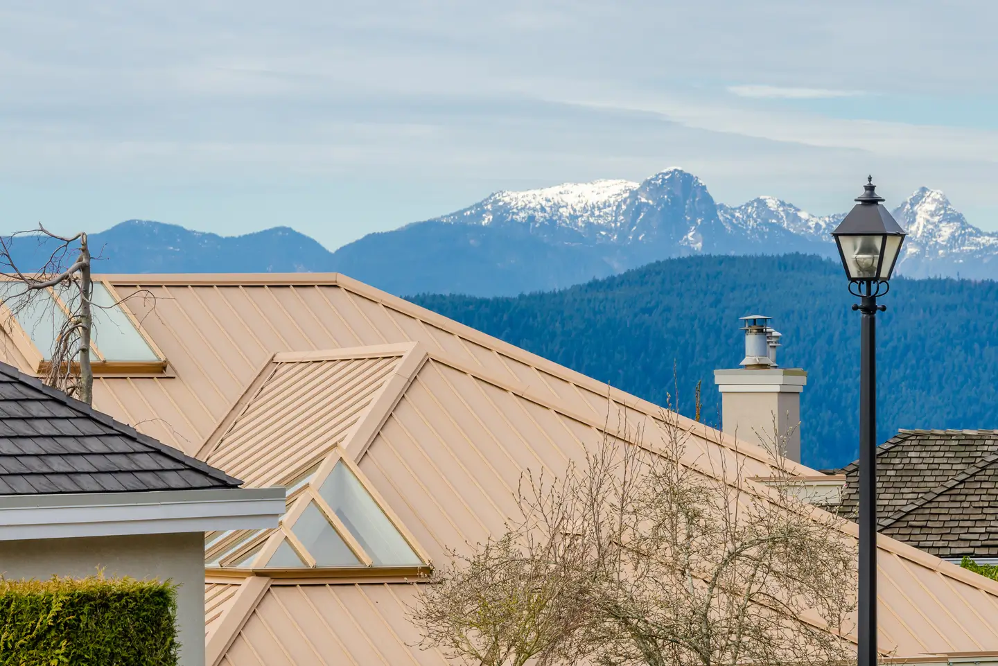 Suburban home rooftop with a beige metal roof, chimney, and mountain backdrop.