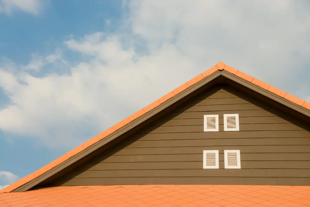 Gable end of a house with brown siding, orange roof tiles, and white vents against a blue sky.